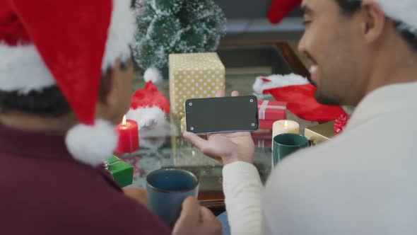 Biracial adult son and senior father in santa hats making smartphone christmas video call