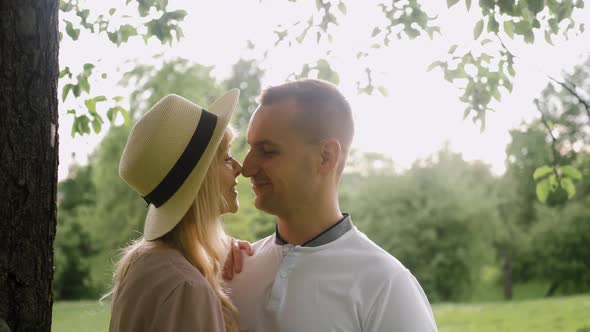 Young Guy with a Woman in a Dress and Hat are Standing By a Tree on a Green Field in Nature in