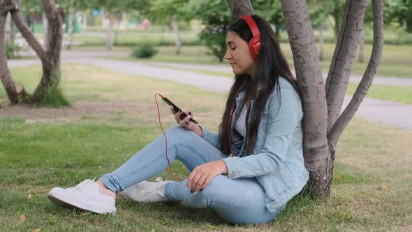 Girl Listening To Music in Headphones Sitting Near a Tree in the Park.