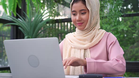 young smiling asian muslim woman working on laptop sitting in living room at home.