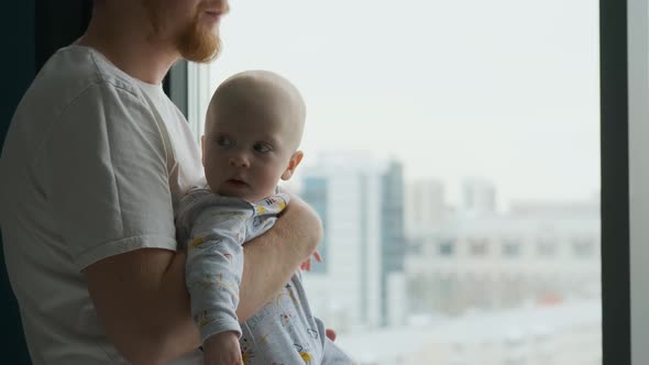 a man holding a curious baby in his arms while standing by the window
