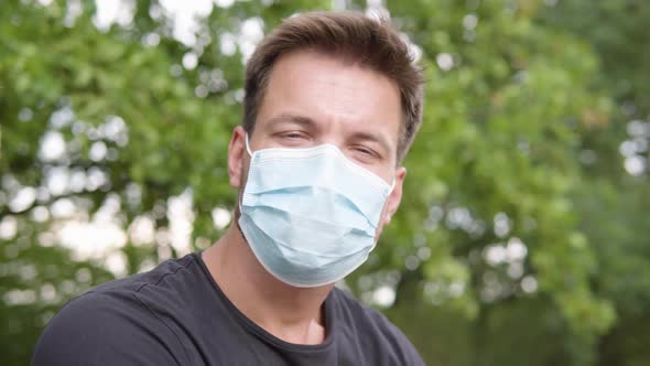 A Young Man Puts on a Face Mask and Looks at the Camera - Closeup - Trees in the Blurry Background