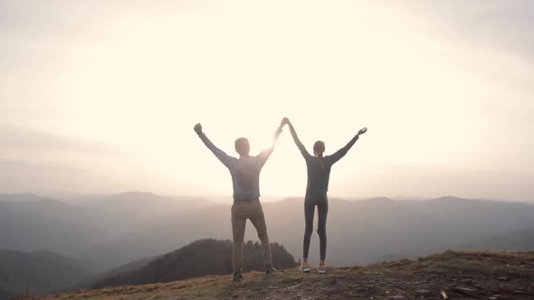 Silhouettes of Couple Hikers Standing Raising Hands on Edge of Mountain Ridge Against Background of