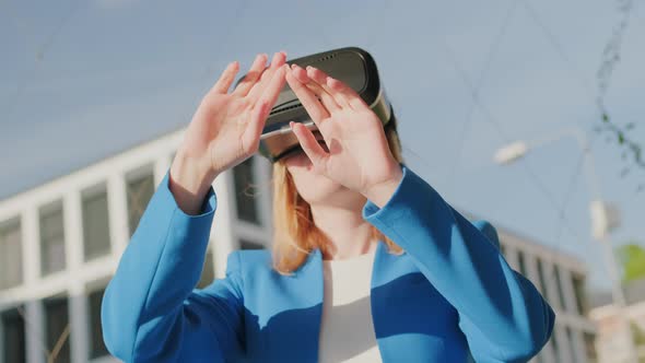 A Happy Woman in the Suit is Standing Outside and Gestures in Augmented Reality in 3D VR Goggles