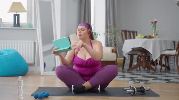 Confident Plussize Caucasian Woman Enjoying Music in Headphones Sitting on Exercise Mat Filming