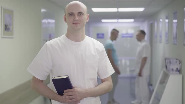 Portrait of Smiling Young Doctor Posing in Hospital Corridor with Two Blurred People Shaking Hands