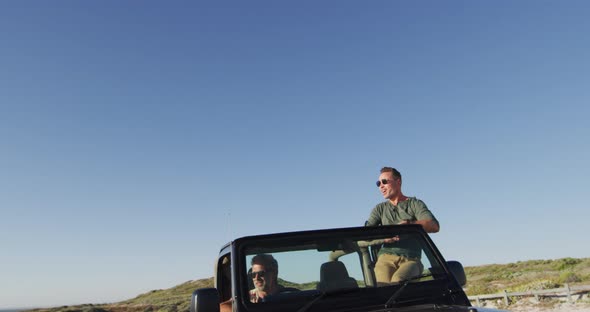 Happy caucasian gay male couple in car admiring the view and waving arms on sunny day at the beach