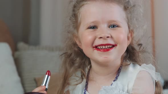 Cheerful little girl cyild paints her lips with bright red lipstick borrowed from her mother.