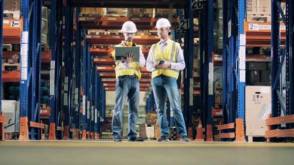 Two Warehouse Workers Talking, Forklift Truck Moving in the Background