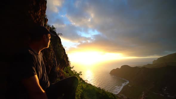 Man Sitting On Mountainside Watching Sunrise Over Sea