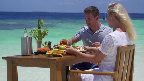 A man and woman eat breakfast on a tropical island beach