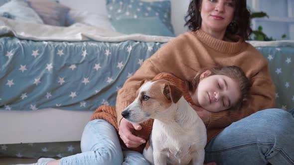 Young Mother and Daughter in Sweaters Sit on the Floor on a Rug with a Dog Jack Russell and