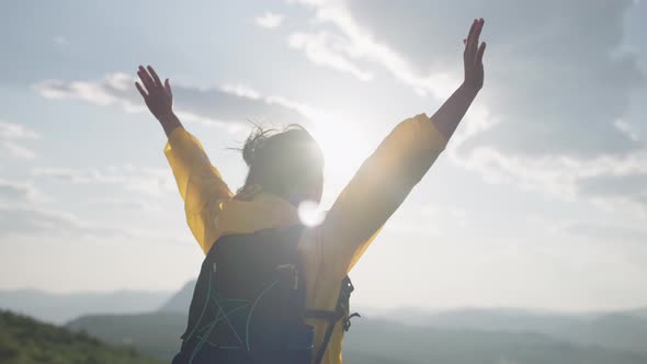 A Happy Girl in a Yellow Jacket Raises Her Hands Uphill on Top of a Mountain