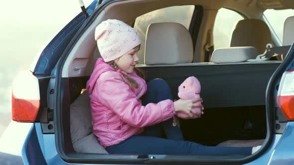 Pretty Happy Child Girl Playing with a Pink Toy Teddy Bear in a Car Trunk