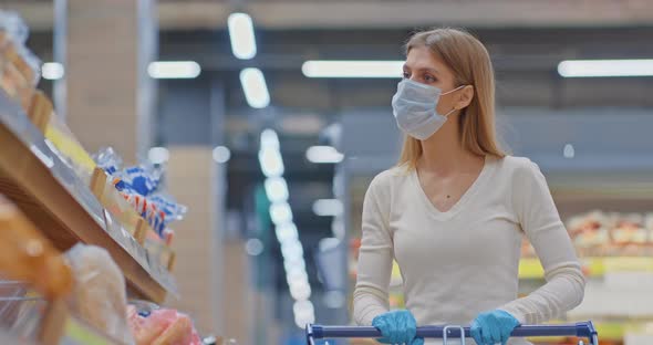 Portrait a Girl in a Medical Mask with a Grocery Cart Approaches the Bread Shelf in the Supermarket