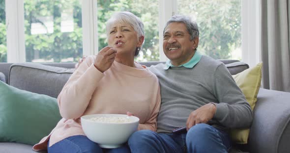 Happy biracial senior couple watching tv with popcorn and having fun at home