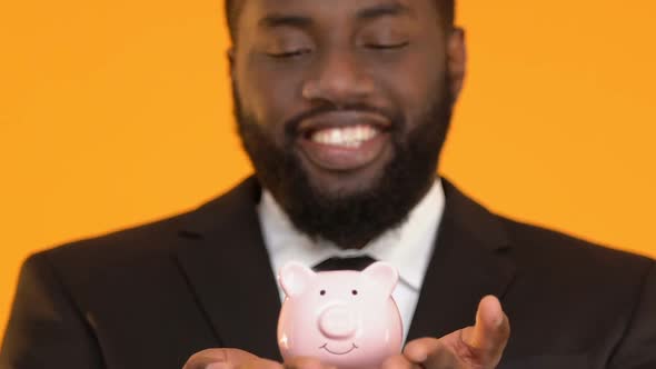 Happy Black Manager in Suit Showing Piggy Bank Into Camera, Savings Concept