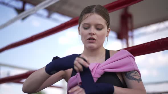 Confident Focused Female Boxer Wrapping Wrist with Boxing Bandage Sitting on Ring Outdoors