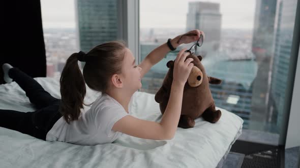 A Cute Girl Lies with Her Favorite Toy on the Bed Near the Window Overlooking the City From Above