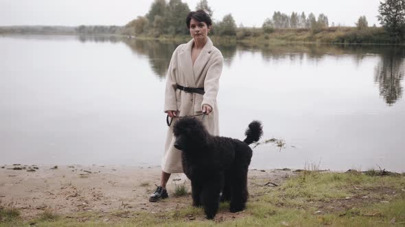 An Exquisite Young Woman in a White Coat Stands on the Shore River and Holds a Large Black Poodle on