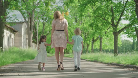 Back View of Young Blond Caucasian Mother Walking with Two Children Along the Alley in Park. Wide