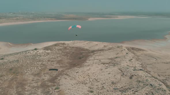 Aerial View Flying Motorised Paraglider Flying Over Arid Coastline Next To Salt Lake. Follow Shot