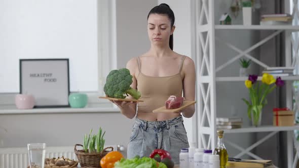 Portrait of Doubtful Young Woman Choosing Between Broccoli and Meat Indoors