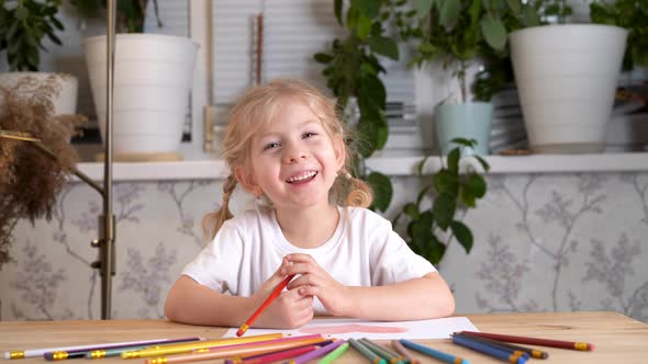 a Small Beautiful Blonde Girl with Gray Eyes and Pigtails Sits at a Table and Smiles at the Camera