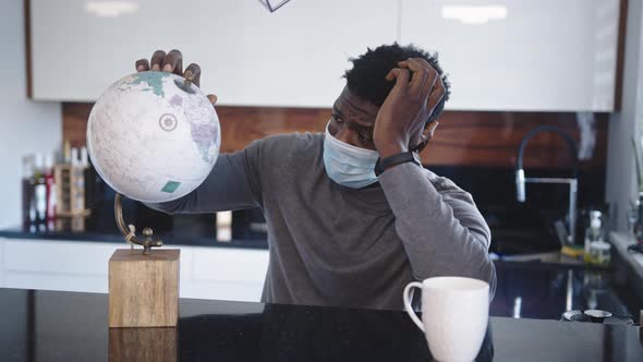 Lonely Black Man Traveler Looking at Globe in His Home with Medical Mask. Traveling During Covid-19