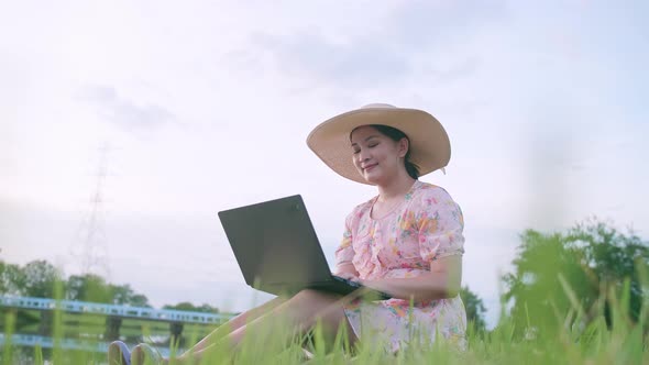 Beautiful Asian woman in hat sitting on lawn working on computer feeling successful.