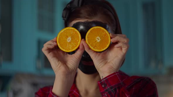30s woman with black cosmetic mask on her face holding orange fruit halves near eyes