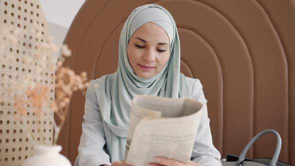 Muslim Woman Wearing Traditional Hijab Reading Newspaper at Table in Cafe