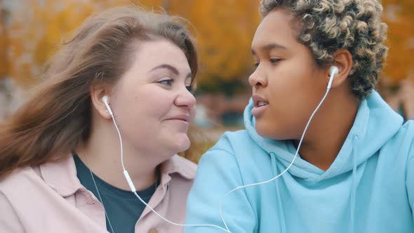 Young Diverse Lesbian Couple Having Fun Listening To Music Sharing Same Earphones in Fall Park