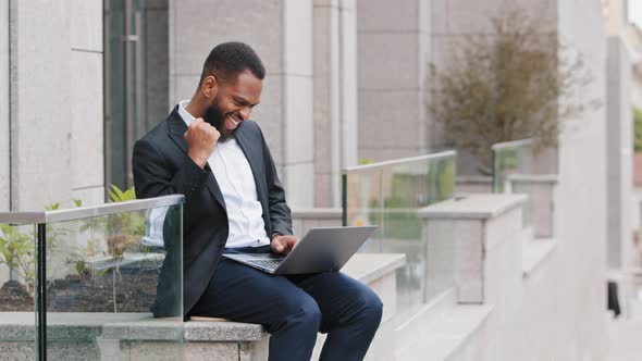 Mixed Race Millennial Man Wearing Blazer Looking at Laptop Screen Outdoors Celebrating Victory After