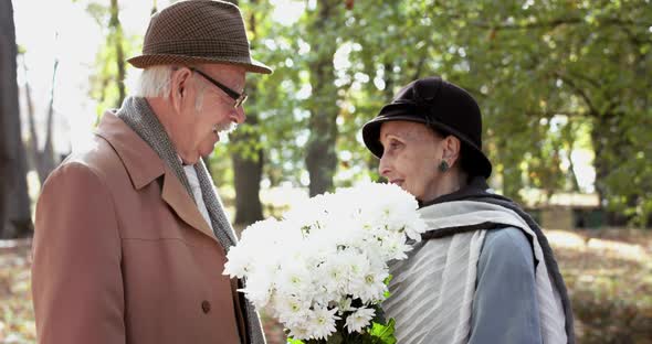 Happy Elderly Woman Plays with Bouquet of Flowers at Her Lovely Cavalier in Park