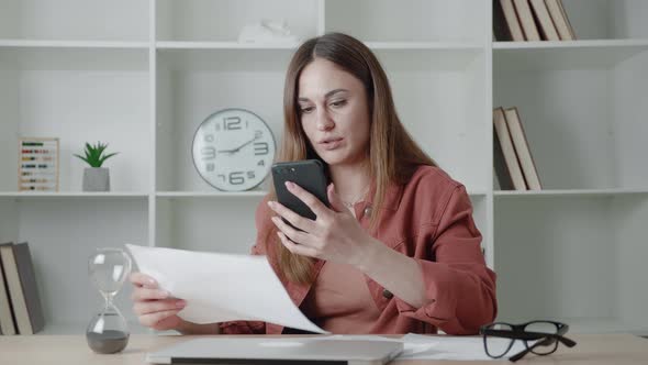 Business Woman Sitting From the Desk Holding Smartphone Have Video Call and Looking Documents