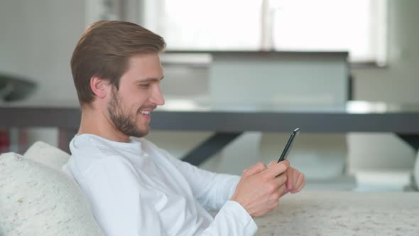 Cheerful Caucasian Man Sits at Home on the Couch and Uses a Smartphone Surfing Internet Pastimes