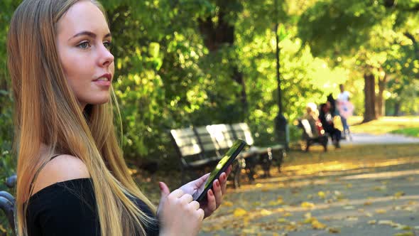 Young Beautiful Woman Sits on the Bench in Park and Works on the Smartphone