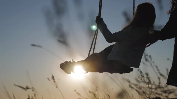 Silhouette of Happy Young Mother and Little Daughter on a Swing at Sun Light