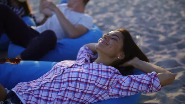 Happy Young Woman Laying on Easy Chair and Smiling Among Group of Friends on the Beach During a