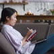 Asian Woman Learning Distantly Using Laptop Computer and Writes Notes While Resting on Sofa at Cosy - VideoHive Item for Sale