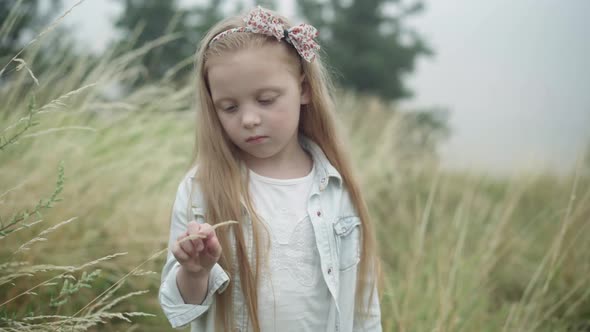 Portrait of Sad Beautiful Caucasian Little Girl Touching Grass Stem Thinking Standing Outdoors on