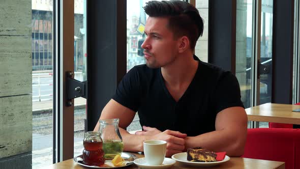 A Young Handsome Man Sits at a Table with Meal in a Cafe, Looks Out a Window and Drinks Tea