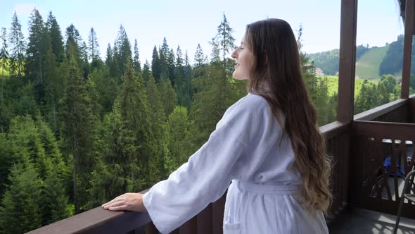 Young Relaxed Woman in Bathrobe Standing on Hotel Room Balcony and Looking on Mountains and Forest