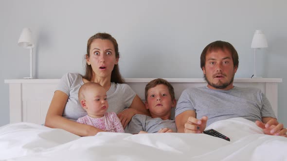 Happy Young European Family with Two Kids Gather on Couch to Watch TV in the Badroom Smiling