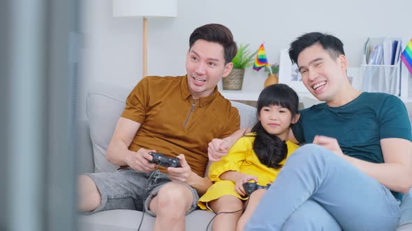 Asian gay couple play video game with young daughter in living room.