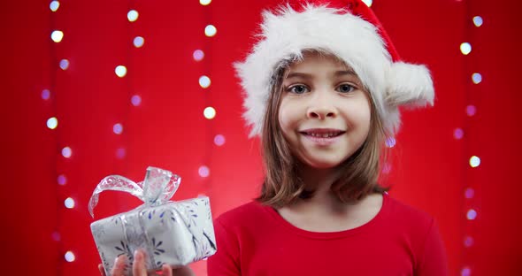 Cute Girl in a Santa Hat Holds a Gift on a Red Christmas Background