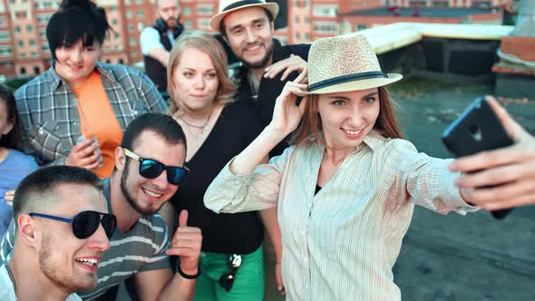 Elegant Young Woman in Hat Taking Selfie Surrounded By Friends Using Smartphone at Rooftop Party