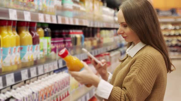 Young Positive Woman Choosing Fresh Orange Juice in a Supermarket Using a Smartphone to Scan a