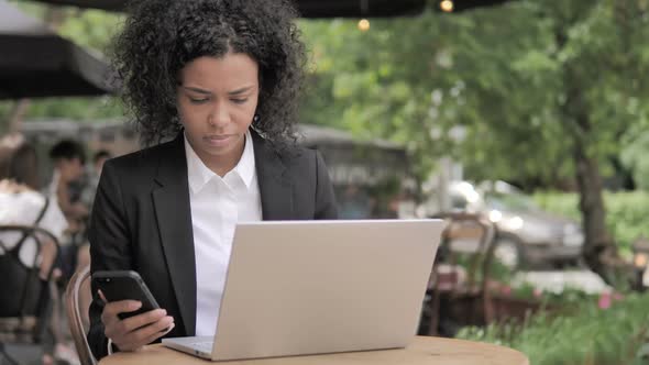 African Woman Using Smartphone and Laptop in Outdoor Cafe